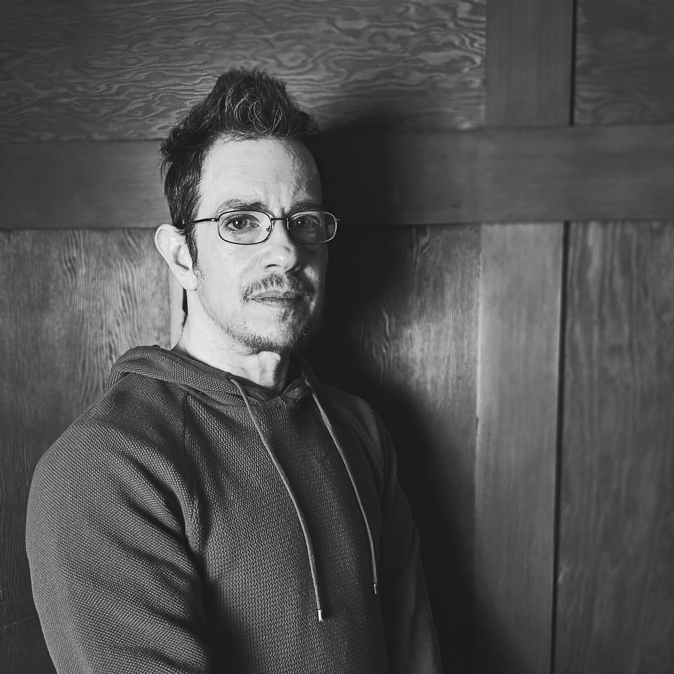 Waist-up portrait shot, Jules is a white person with short brown hair and a goatee, he is wearing thin black glasses and a dark hoodie. He is looking at the camera without smiling. Photo credit: Jules Sherred.