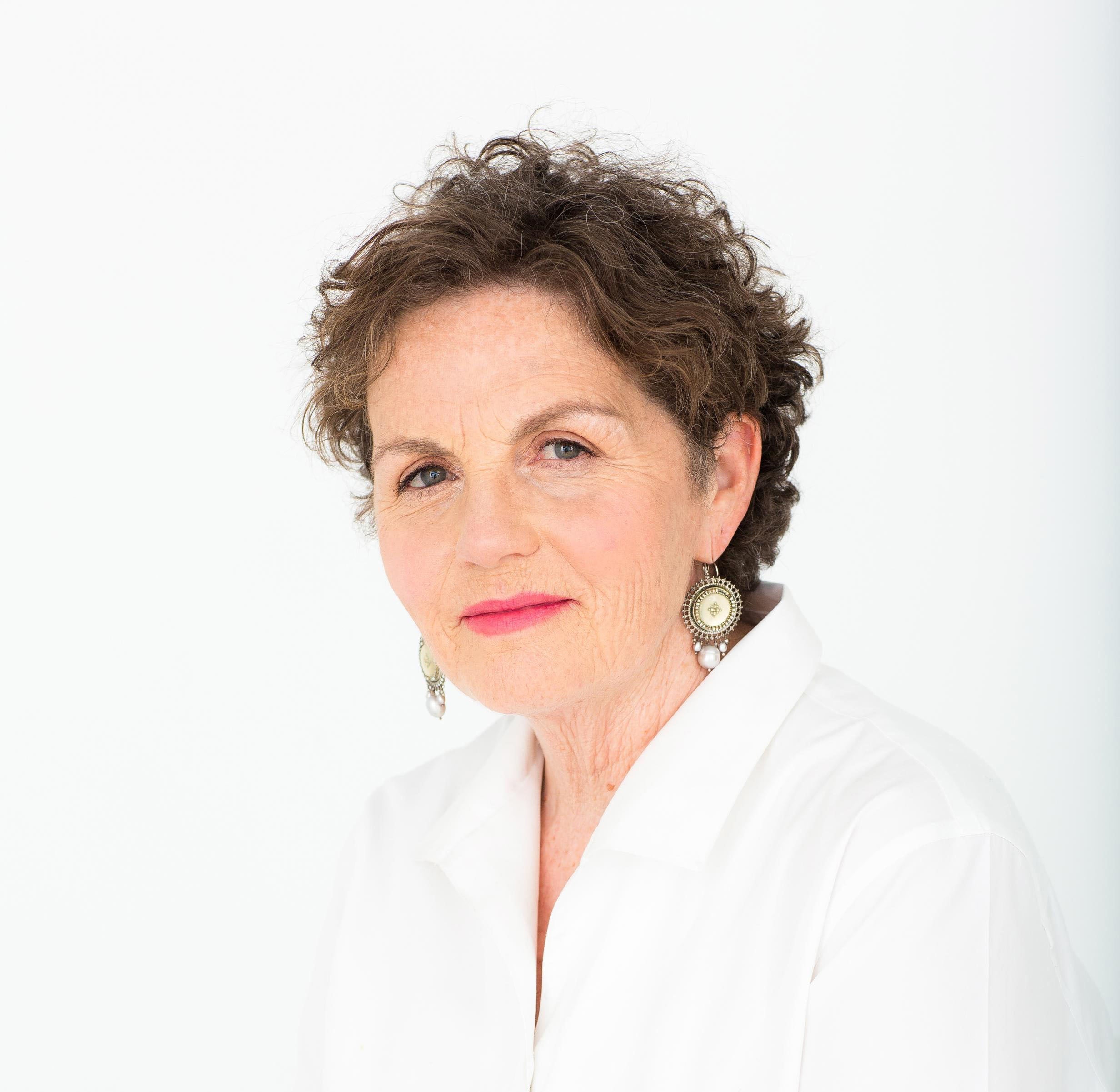 Shoulder-up portrait shot, Iona is a white woman with short brown wavy hair., wearing a white collared shirt, light pink lipstick, and gold earrings. She is looking at the camera. Photo credit: Anick Violette.