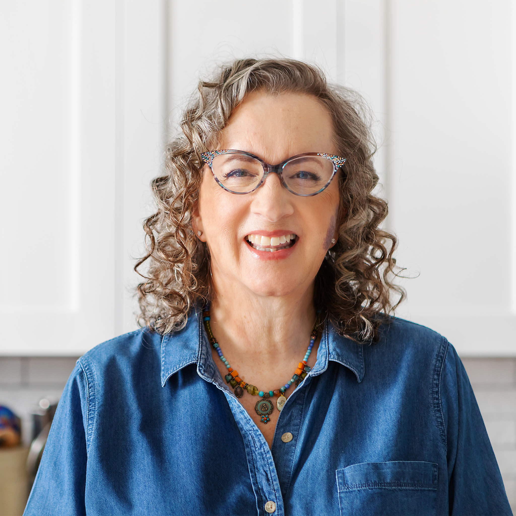 Torso-up portrait shot, Karen is a white woman with blonde-grey curly shoulder-length hair, wearing wing-tipped glasses, a chunky necklace and denim button-down, she is smiling toothily and looking at the camera. Photo credit: Dana Pugh.