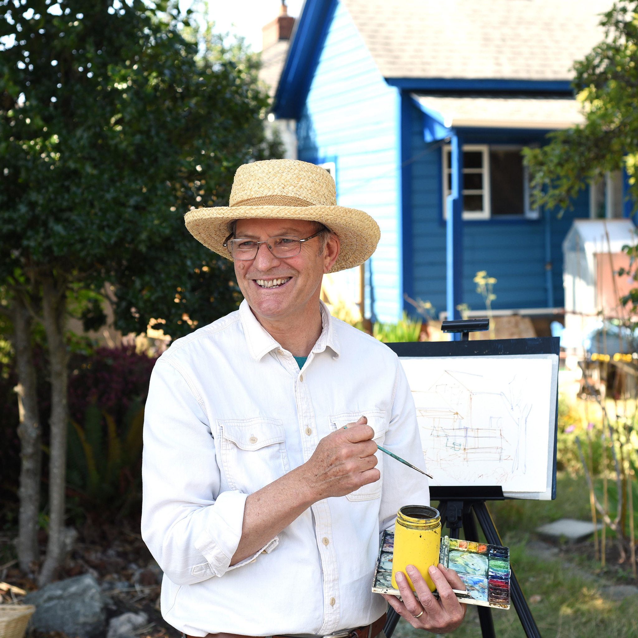 Waist-up portrait shot, Robert is a white man wearing a straw hat, glasses, a white button-down. He is standing in front of an easel with paintbrush in hand and paint in the other, smiling looking off in the distance. Photo credit: Don Denton.