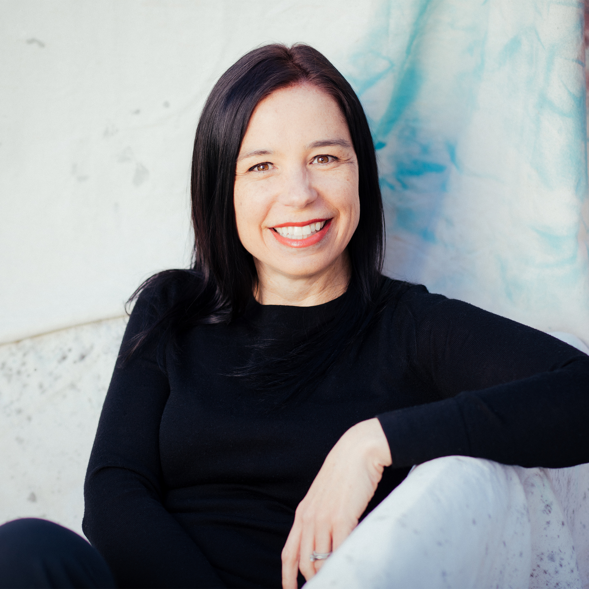 waist-up portrait shot, Ann is a white woman with straight dark hair, wearing a black shirt, she is sitting in a white chair, looking at the camera smiling. Photo credit Jennifer Rowsom.