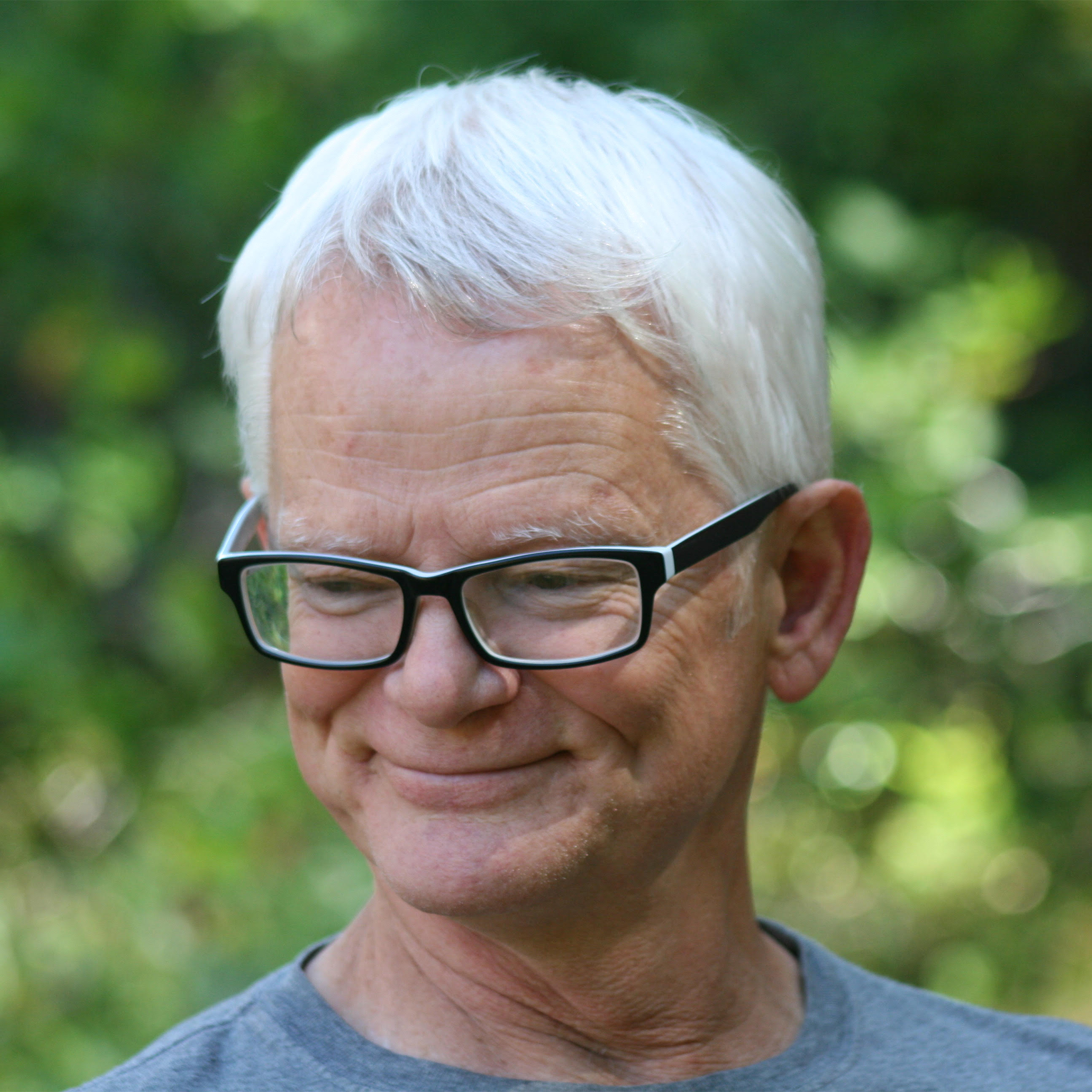 Headshot portrait, Grant is a white man with white hair, wearing thick black rimmed glasses and a grey t-shirt, he is looking down and away from the camera smiling. Photo credit: Eden Buday.