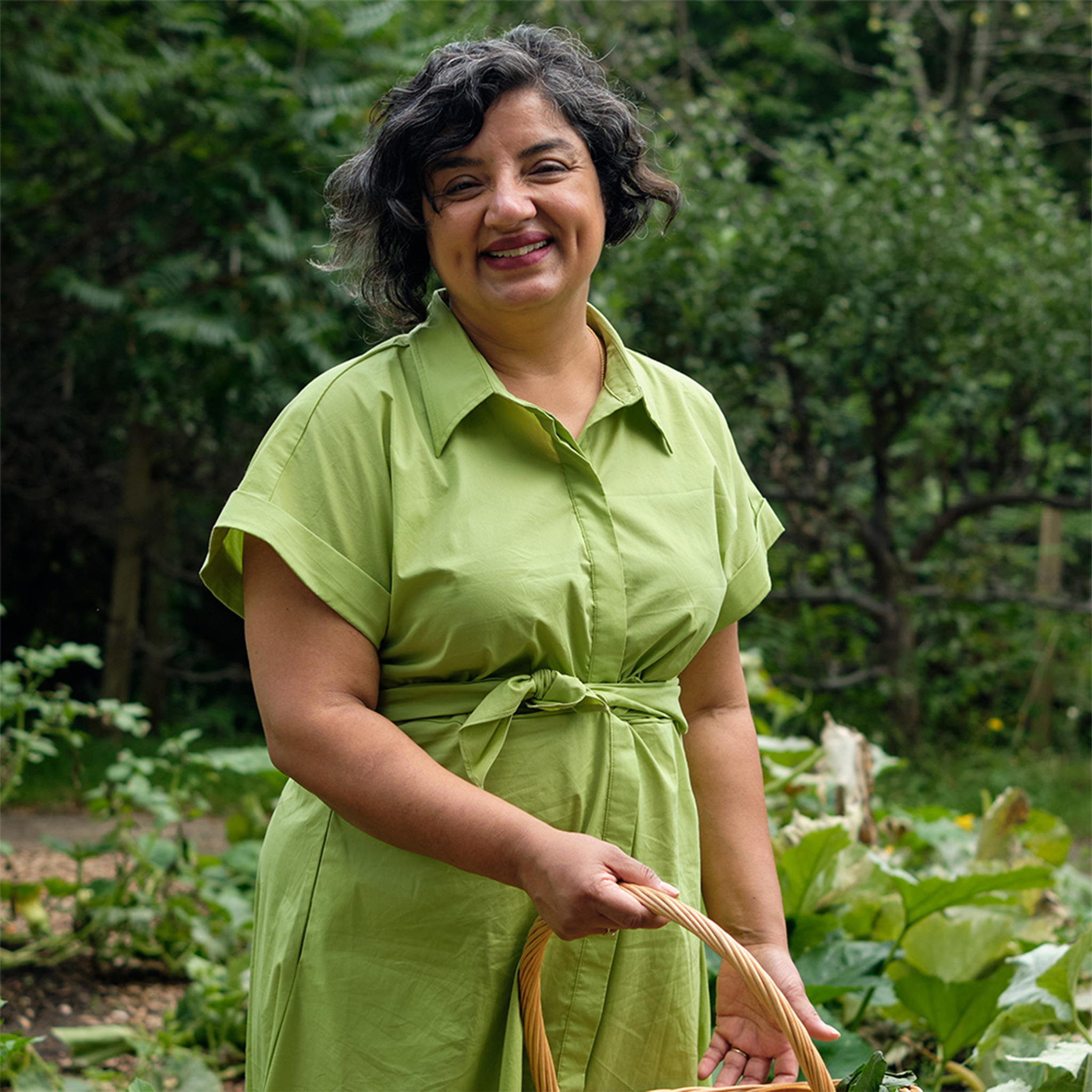 Full body portrait, Puneeta is a brown Indian woman with short dark wavy hair, wearing a green wrap dress. She is standing in the garden, holding a basket and looking at the camera smiling. Photo credit: Diana Muresan.