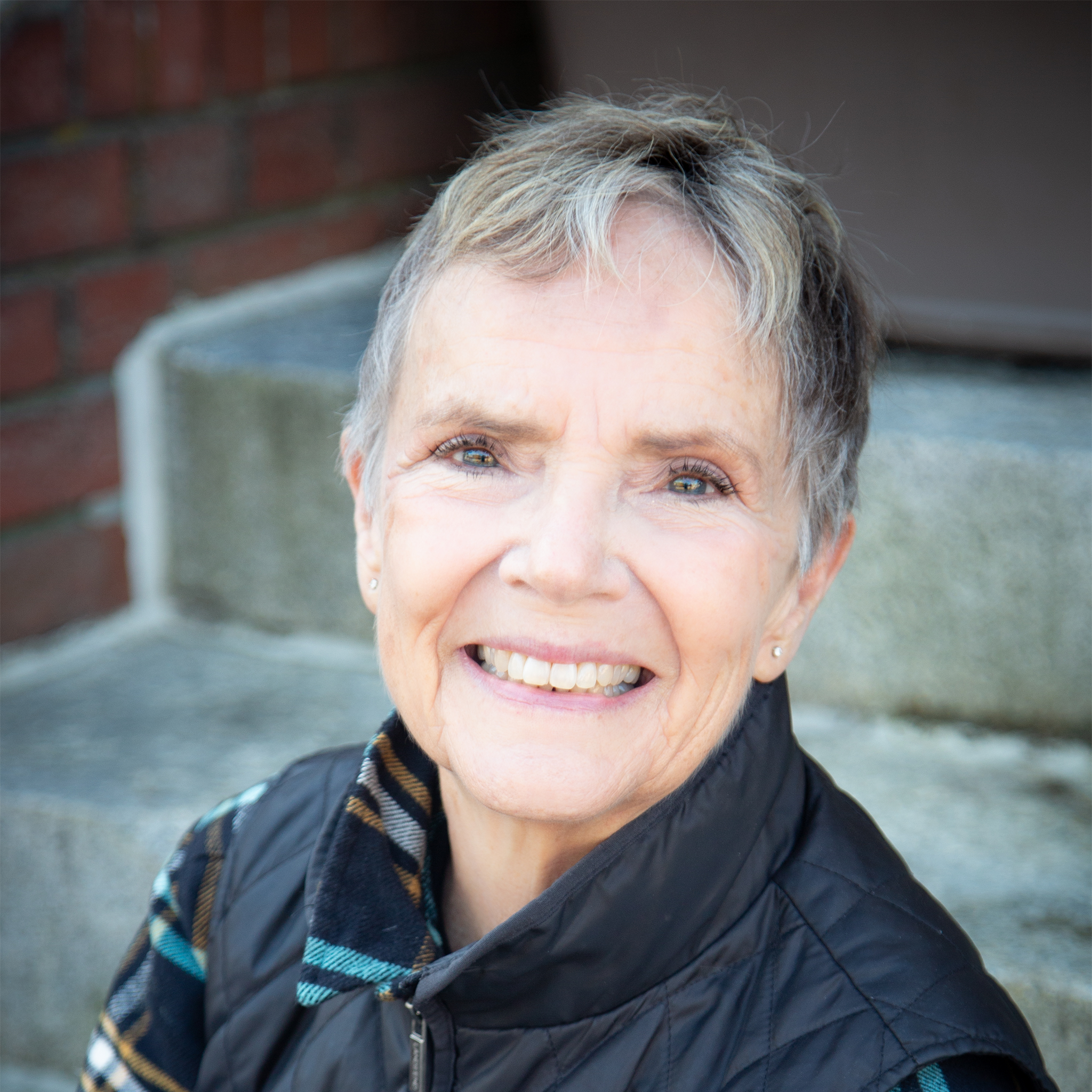 shoulder-up portrait, Leslie is a white woman with short salt and pepper hair, wearing a black fall vest and stud earrings, she is looking up at the camera and smiling large. Photo credit: Sarah Mickel Photography.