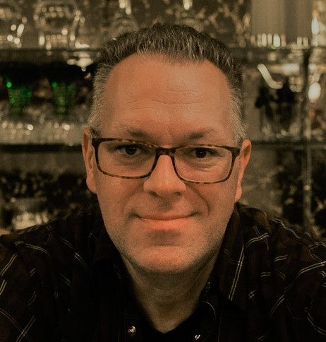 Shoulder-up portrait, Ian is a white man with salt and pepper hair, wearing thin tortoise rimmed glasses and a black collared shirt. He is looking at the camera smirking. Photo credit: Ian Gibbs.