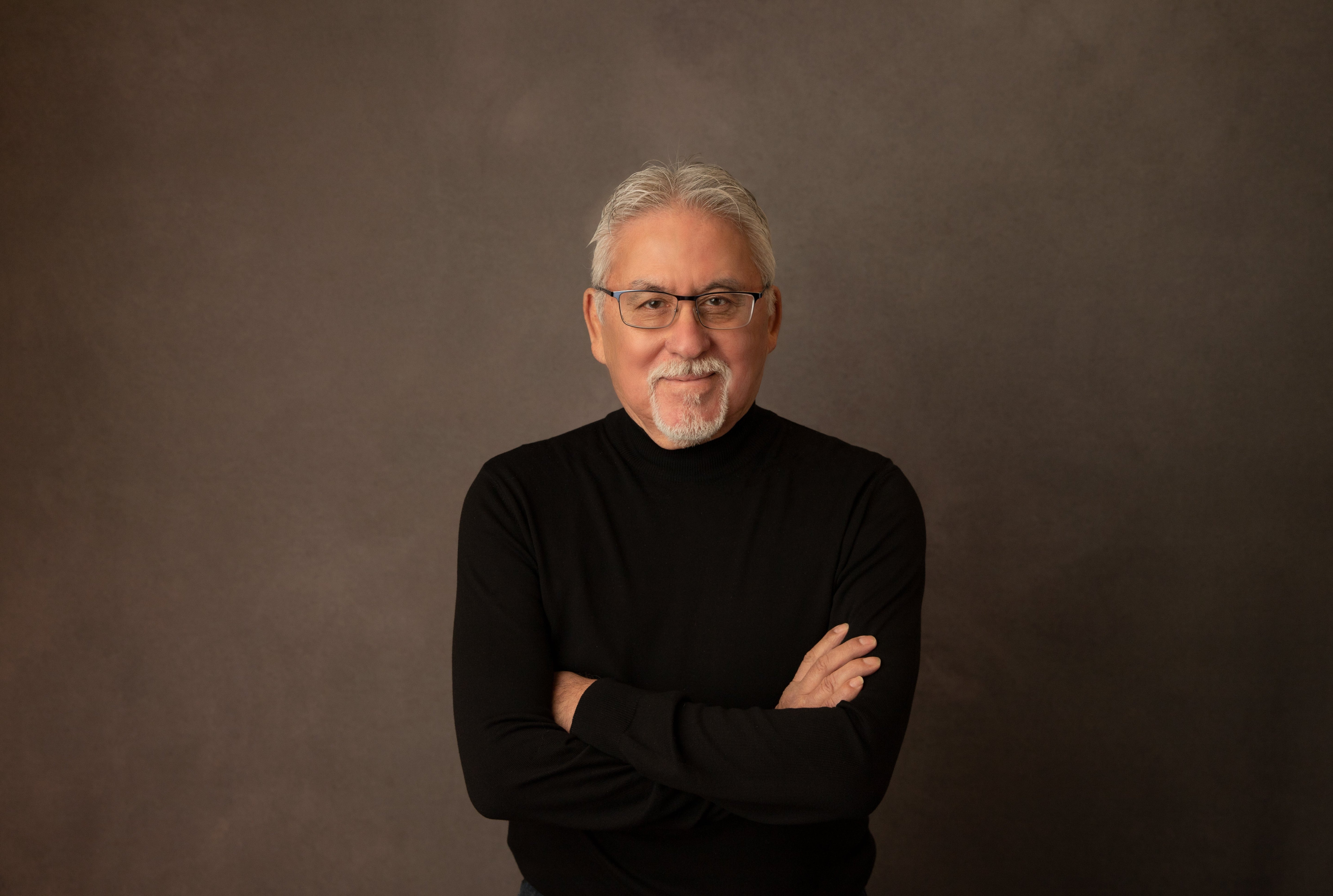 Waist-up portrait shot, Brian is an Indigenous person with grey hair parted in the middle, wearing glasses, and a black turtleneck sweater. His arms are crossed and he is looking into the camera smirking. Photo credit: Fiona Blaquier.