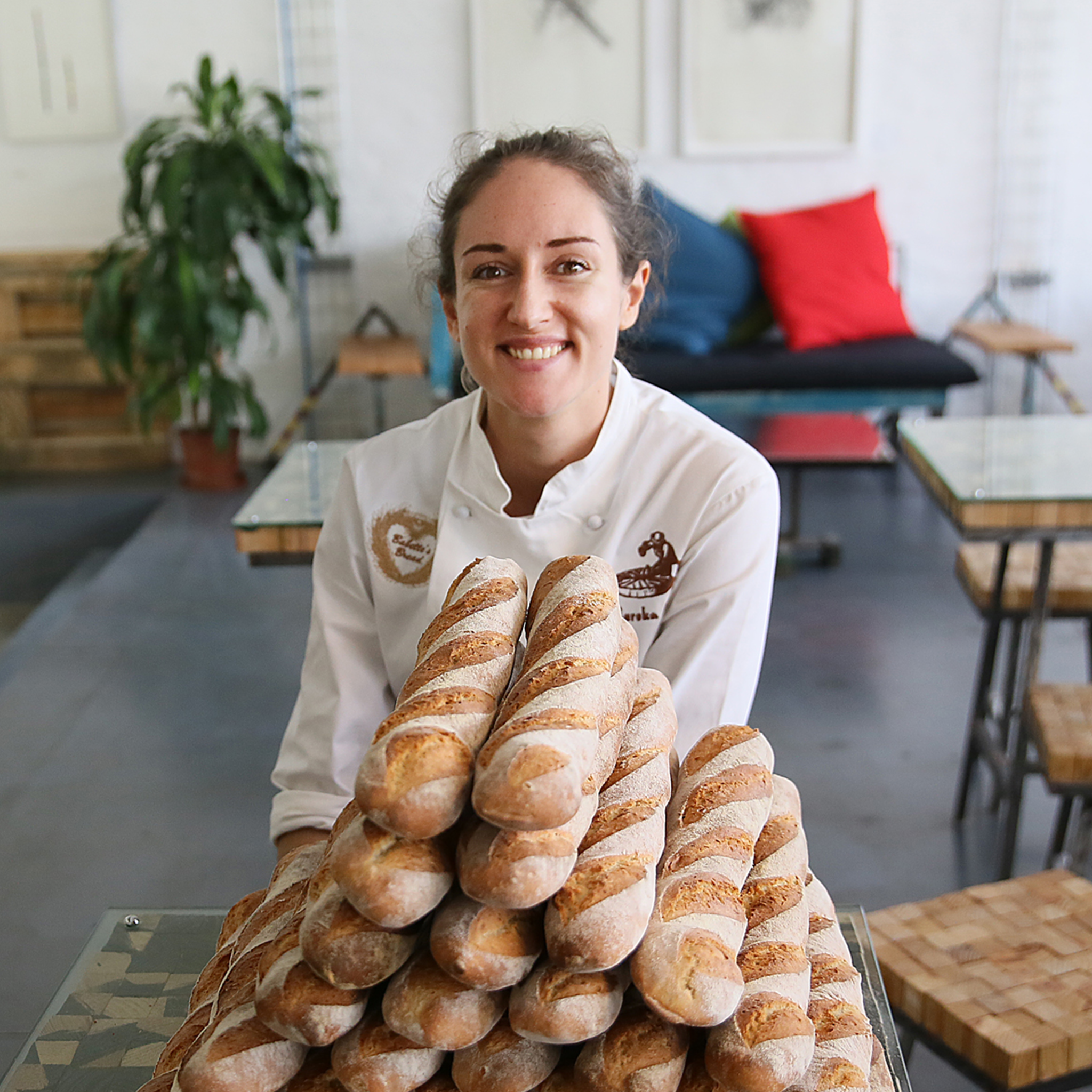 Torso-up portrait shot, Babette is a white woman with dark hair pulled back into a bun, wearing a white chefs jacket she is leaning on a table stacked with loaves of baguettes. She is looking at the camera smiling. Photo credit: Alon Skuy.