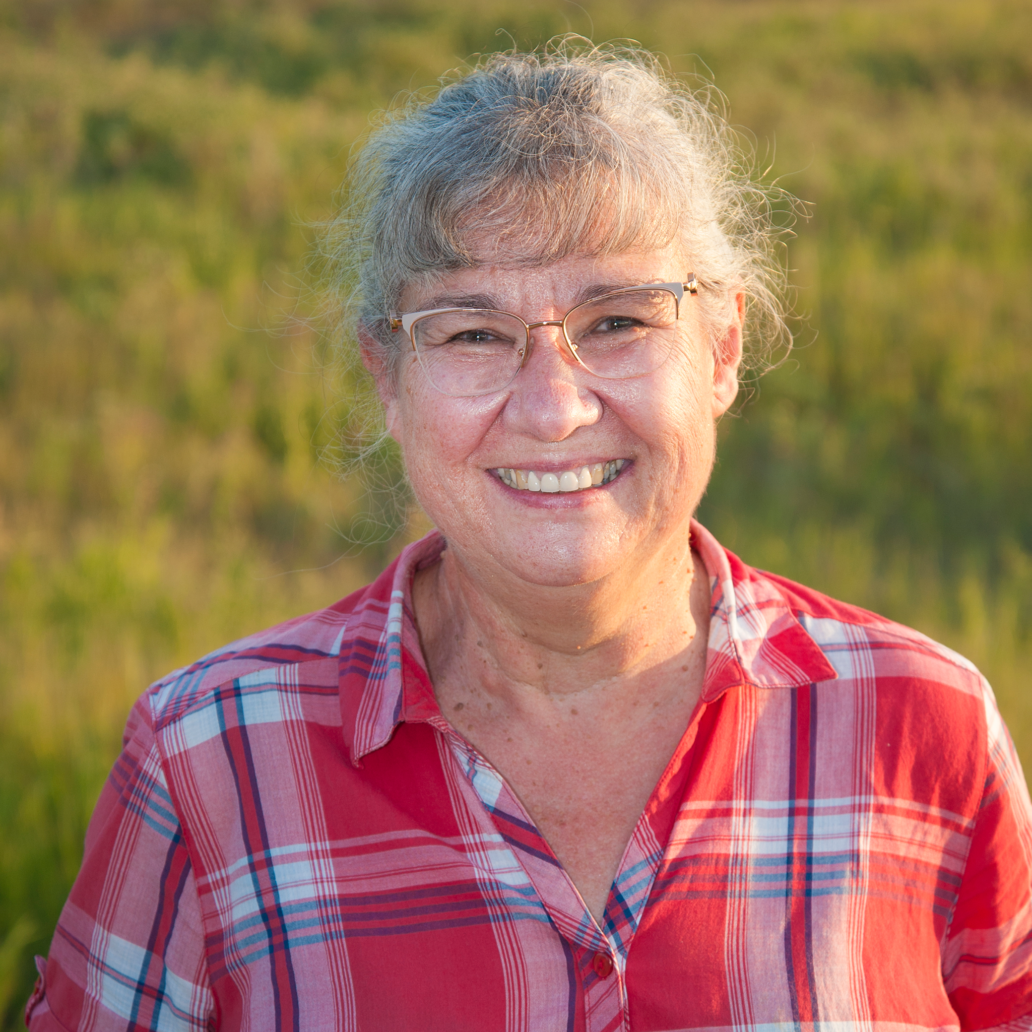 Shoulder-up portrait shot, Janet is a white woman with grey hair with bangs and hair pulled back, wearing silver rimmed glasses and a red plaid shirt, she is looking at the camera and smiling. Photo credit: Steve Melrose.