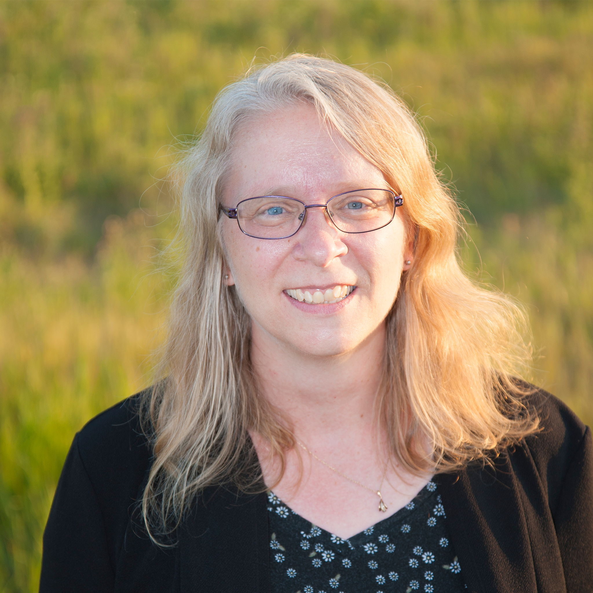 Shoulder-up portrait shot, Sheryl is a white woman with shoulder length blonde hair, wearing thin black rimmed glasses, a black cardigan and patterned shirt, she is looking at the camera smiling. Photo credit: Steve Melrose.