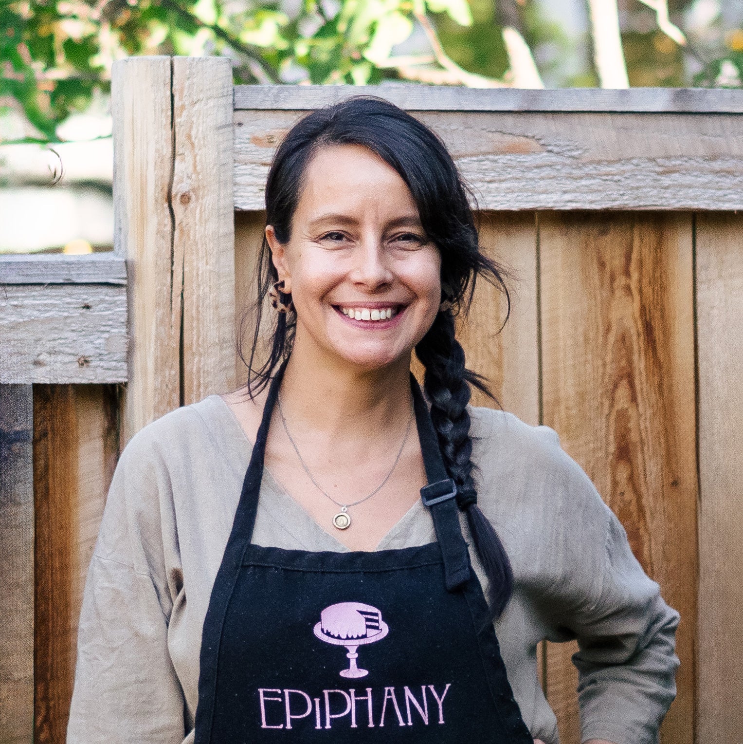 Torso-up portrait shot, Melissa is a woman with dark braided hair, wearing a tan v-neck shirt, necklace, and black apron that says Epiphany. She is looking at the camera smiling.