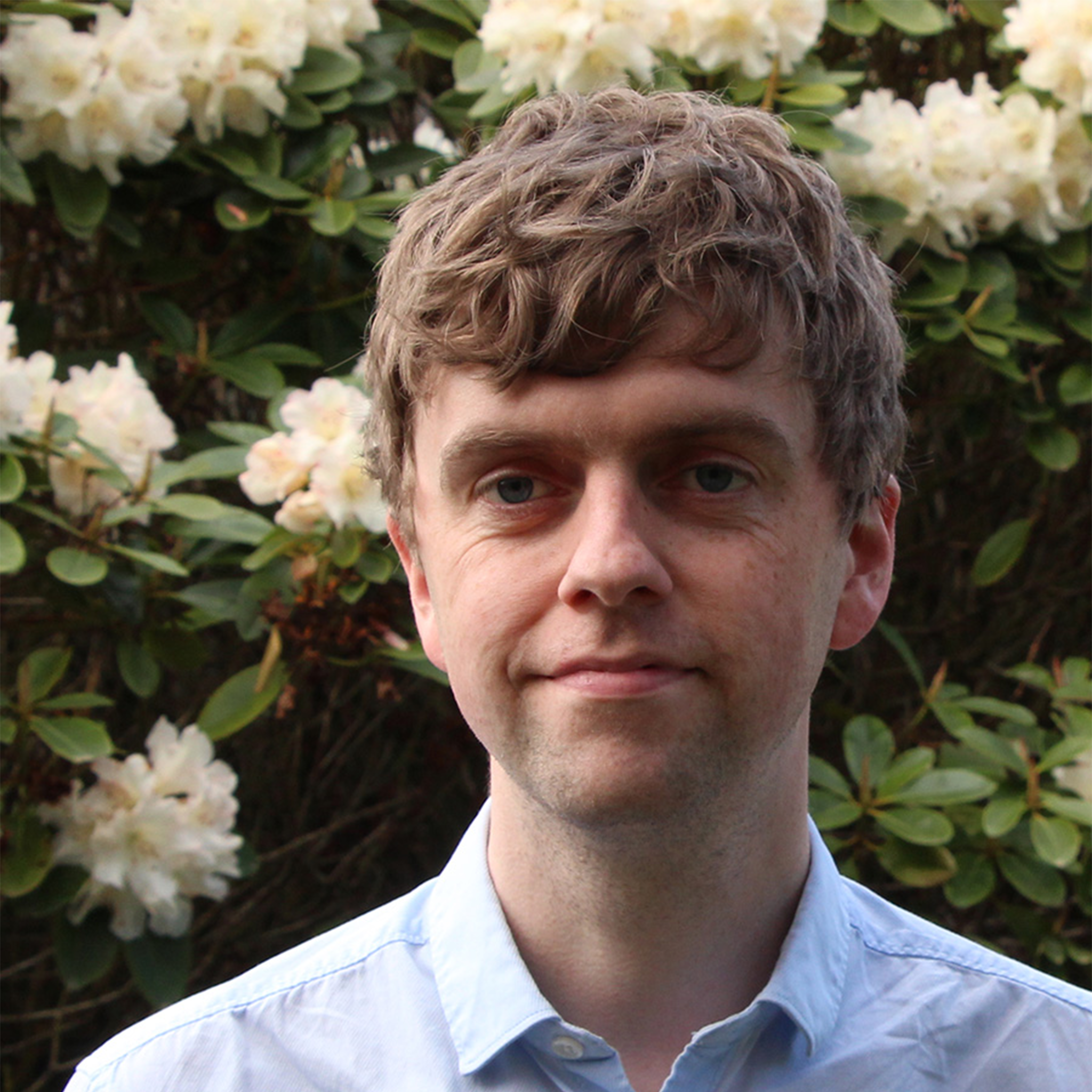 Shoulder-up portrait shot, Sam is a white man with short light brown hair, wearing a light blue button up, he is standing in front of a wall of flowers, looking past the camera unsmiling. Photo credit: Cliff Haman.