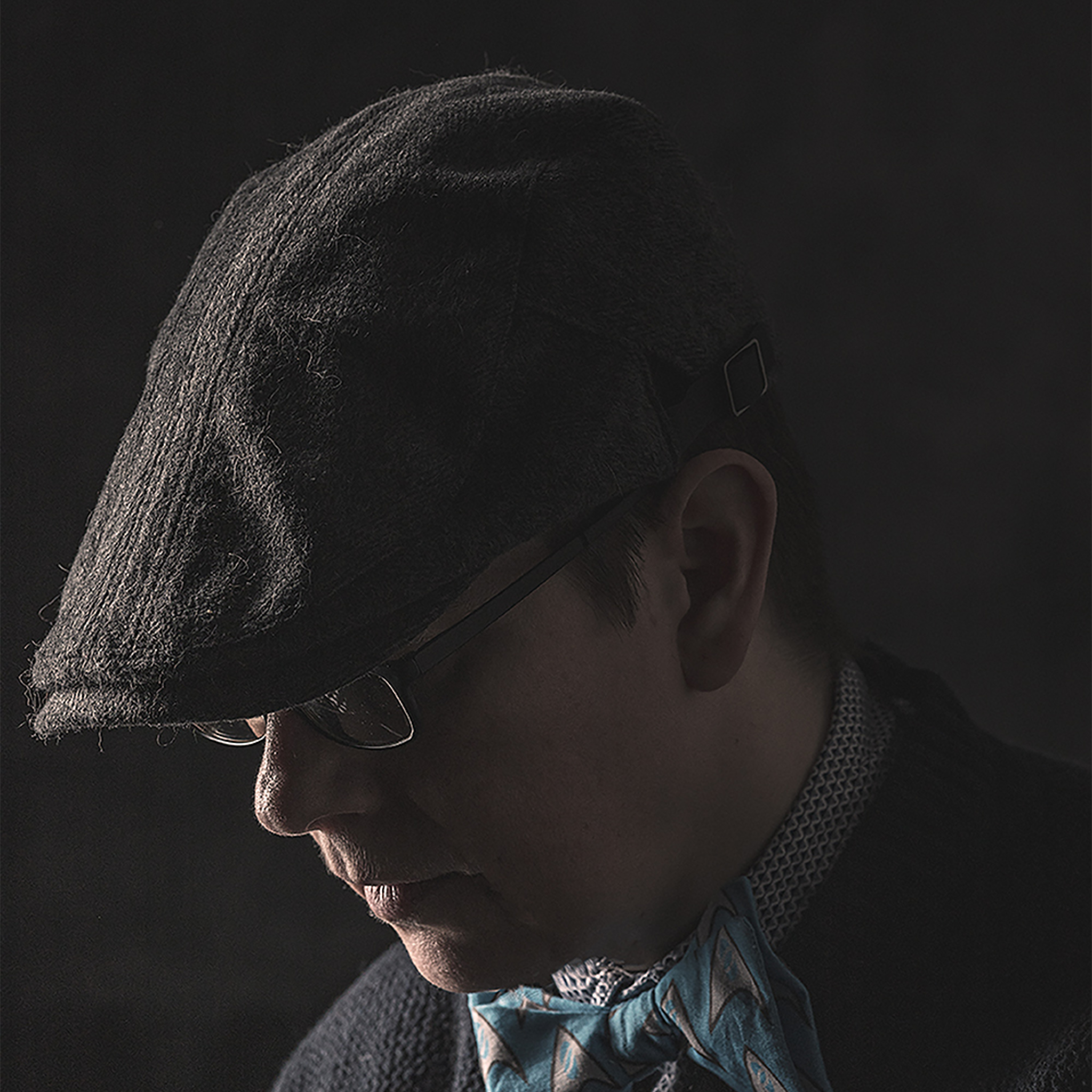 Shoulder-up portrait shot, Jules is a white person with brown hair, wearing thin black glasses, a black newsboy cap, blue and grey Star Trek bowtie, grey patterned collared shirt, and knitted navy-blue zip up cardigan. He is looking down without smiling. 