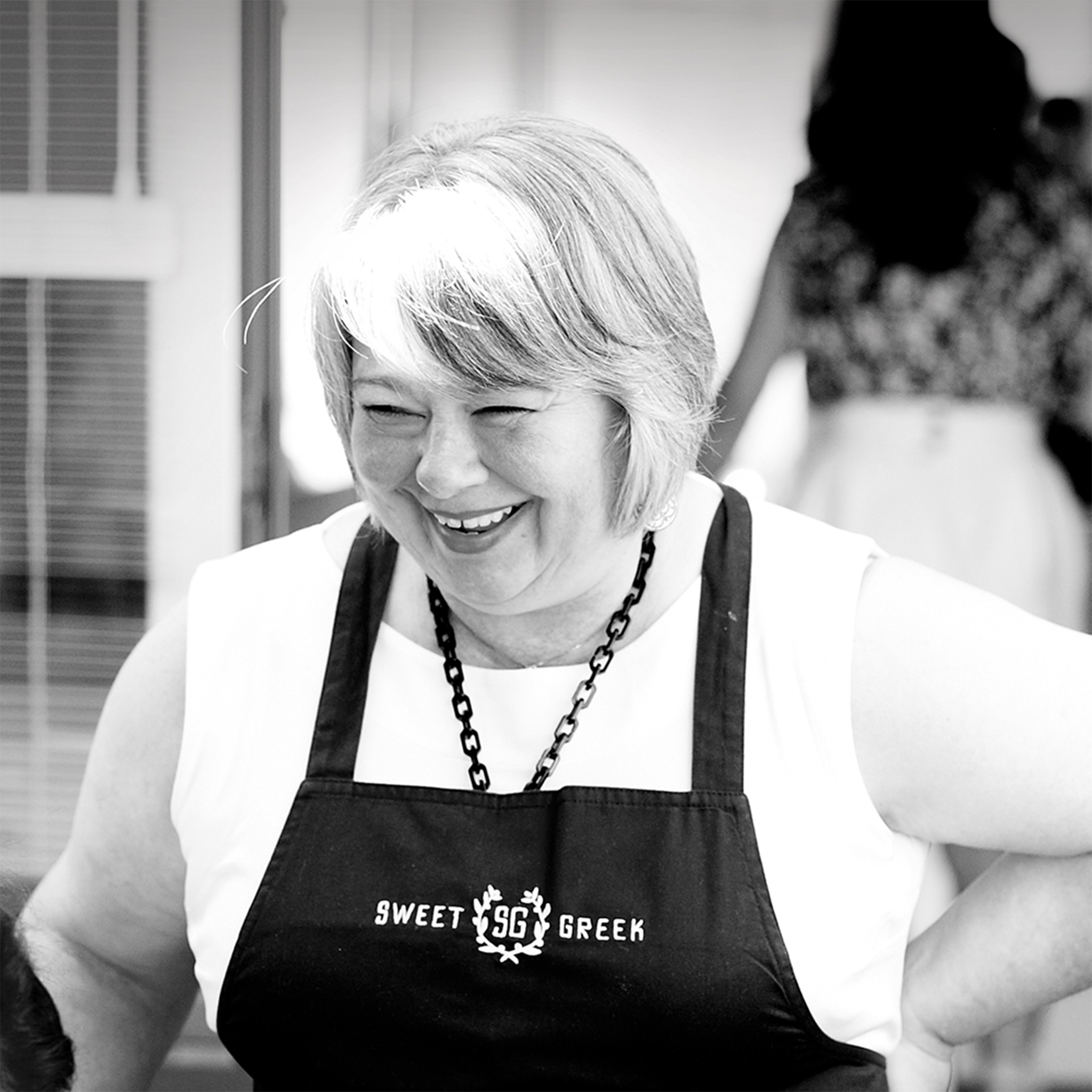 black and white photo, waist-up portrait, Kathy is a white woman with grey short hair, wearing a white shirt under her apron that reads sweet greek, one hand on her hip she is looking at a customer smiling toothily.