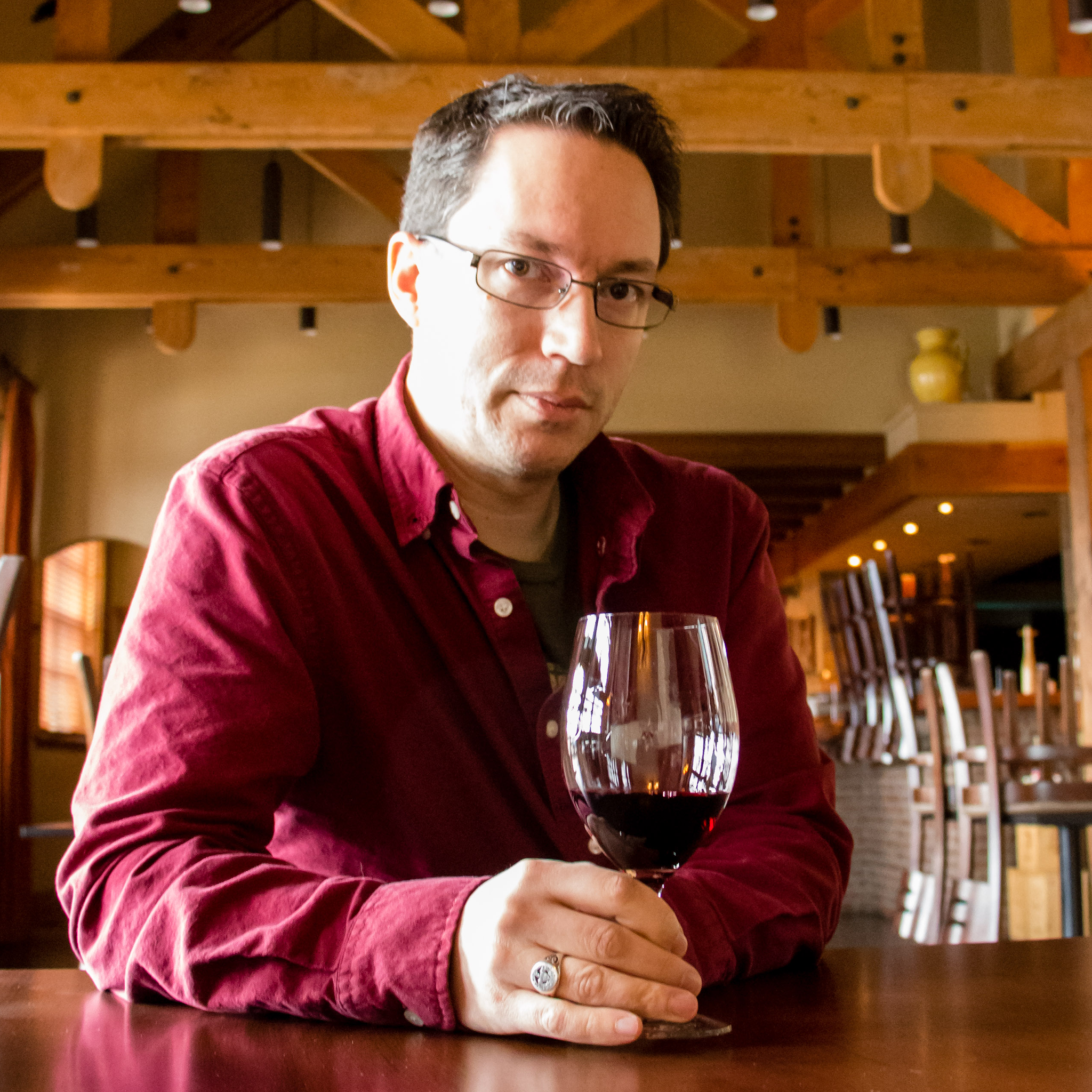 Waist-up portrait photo, Luke is a white man with short brown hair, wearing thin rimmed glasses and a burgundy button up shirt. He is leaning on a bar top with a glass on red wine in hand, looking at the camera unsmiling. Photo credit: Jen Jensen.