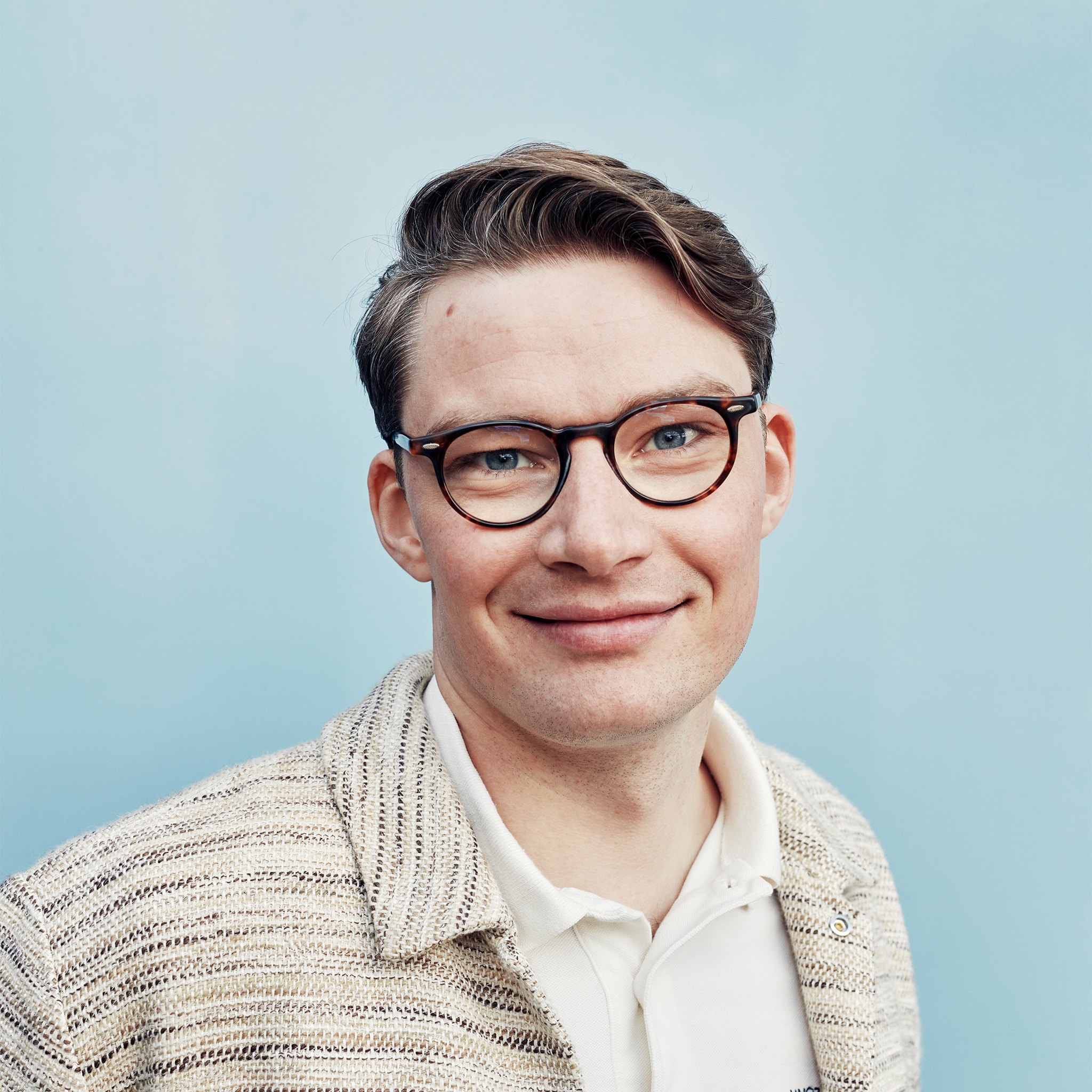 Shoulder-up portrait photo, Rasmus is a white Swedish man with brown hair parted to the side, wearing round brown glasses, a cream sweater over a white polo. He is looking directly at the camera smiling. Photo credit: Andreas Houmann.